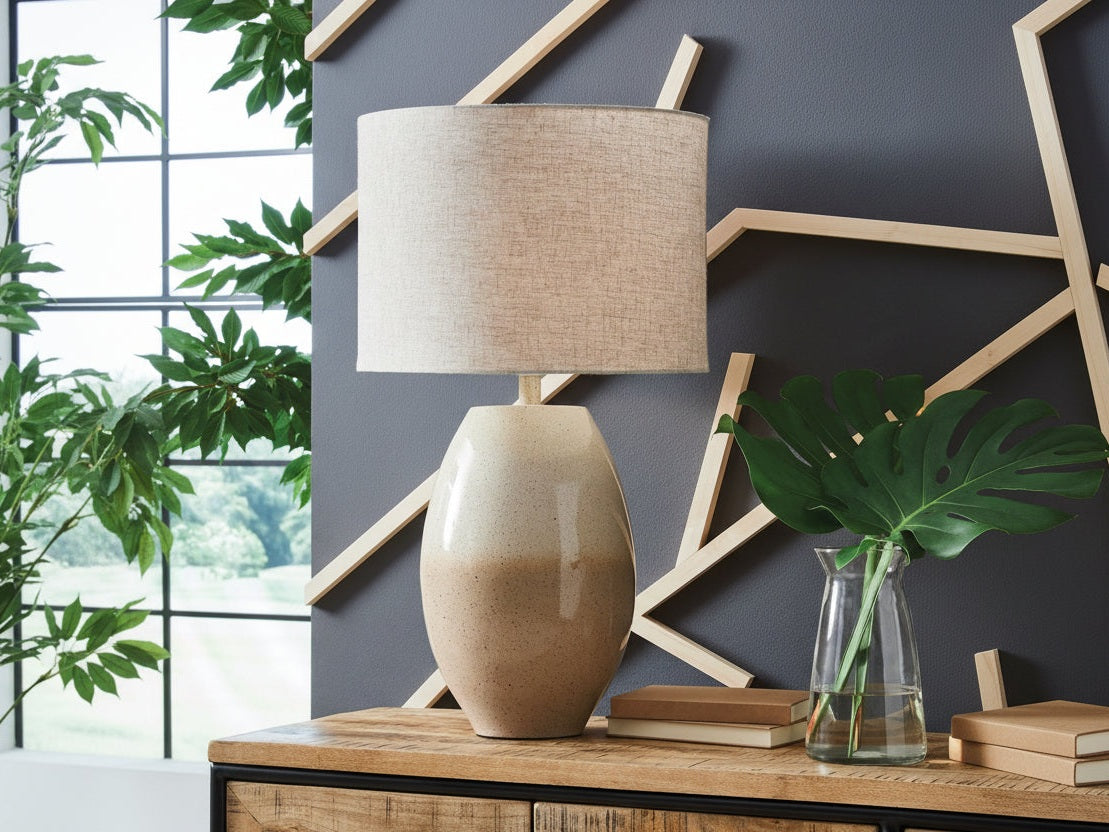 The Linas beige table lamp on a wooden surface with a gray wall and plant in the background.