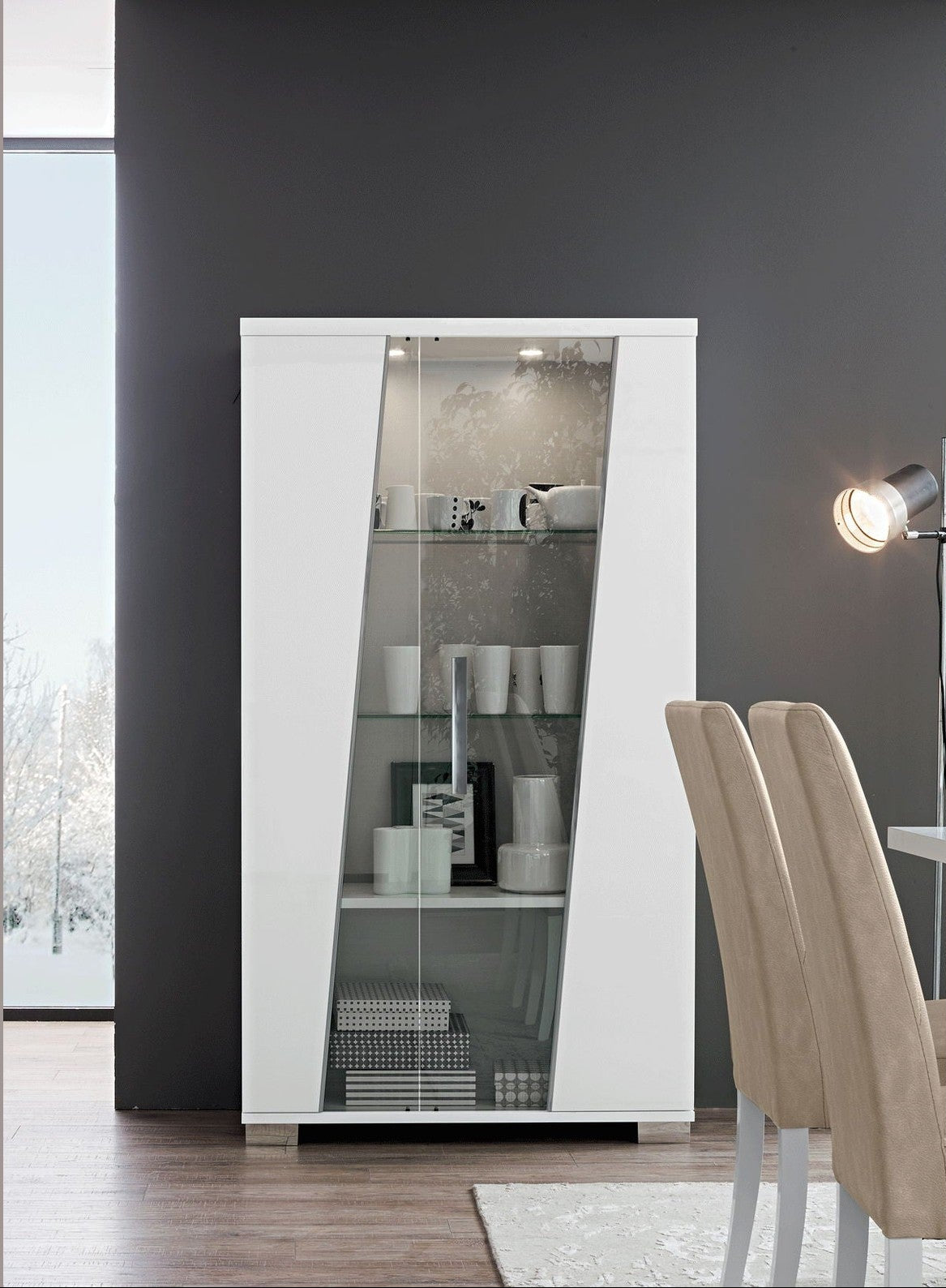 A tall white glass-front display cabinet with interior shelving, shown in a modern dining room setting next to beige upholstered chairs and a floor lamp.