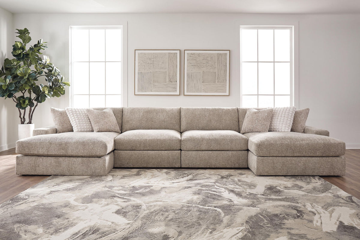 A light beige upholstered sectional sofa with a left chaise and matching accent pillows, shown in a bright, modern living room with a large gray and white patterned rug.
