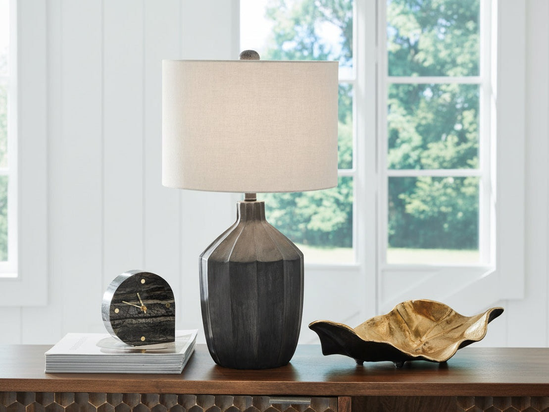 Jilton table lamp with a beige shade on a wooden surface near a window with greenery outside.