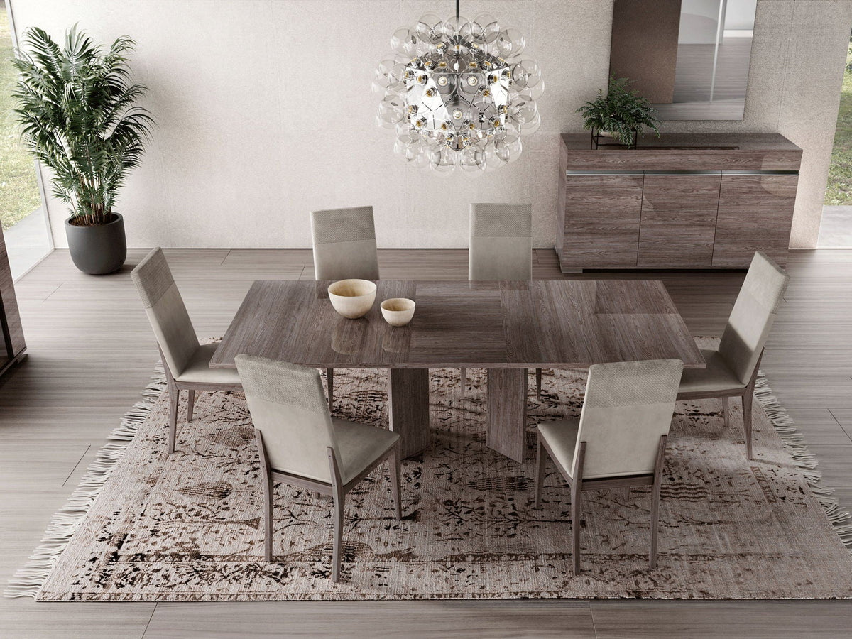 A modern dining room set featuring a rectangular wood-look table, six upholstered dining chairs, a patterned area rug, and a decorative glass chandelier hanging above.