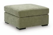 A square upholstered ottoman with a textured fabric in a muted sage green color, featuring black block feet, shown on a white background.