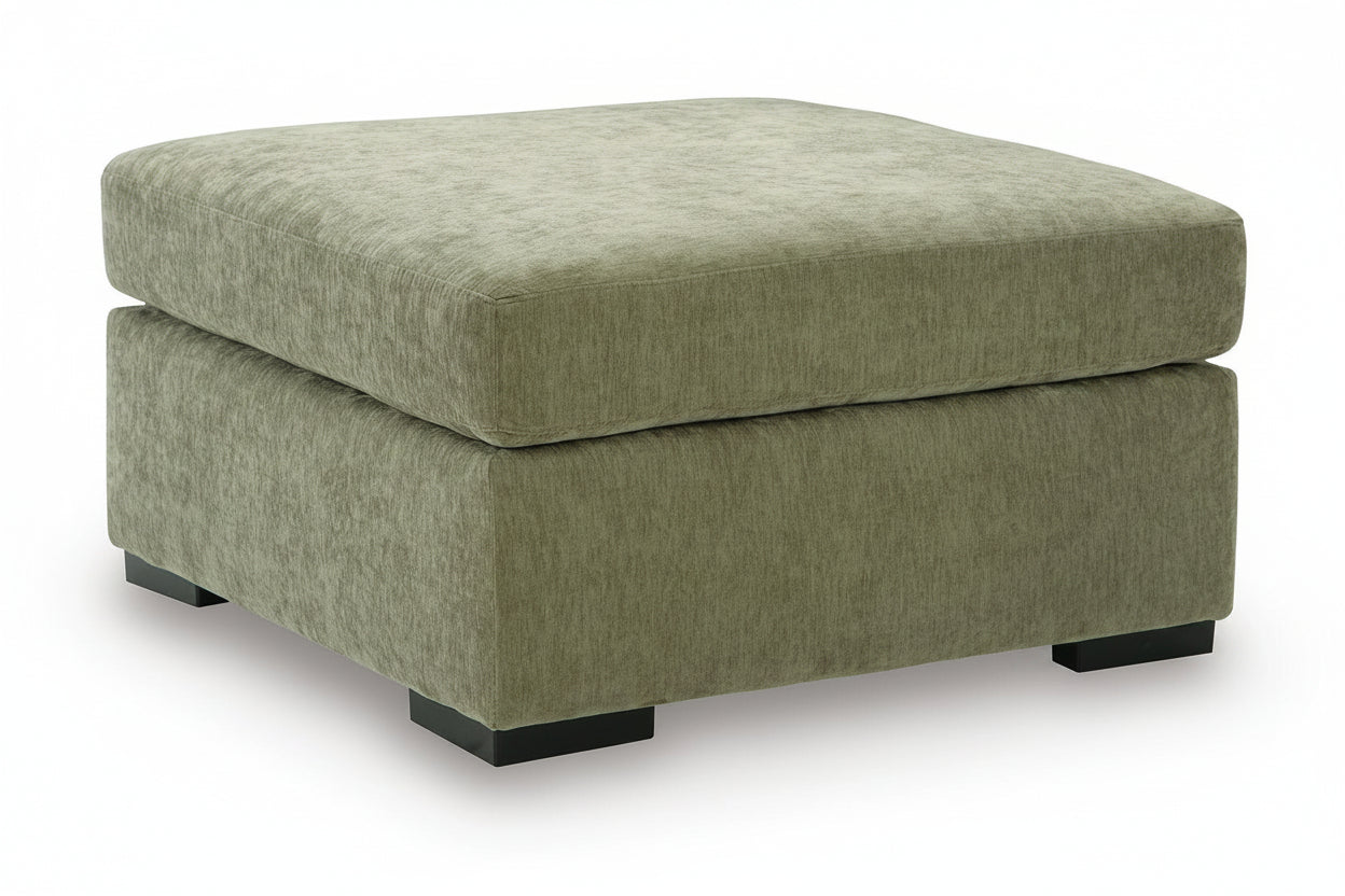 A square upholstered ottoman with a textured fabric in a muted sage green color, featuring black block feet, shown on a white background.