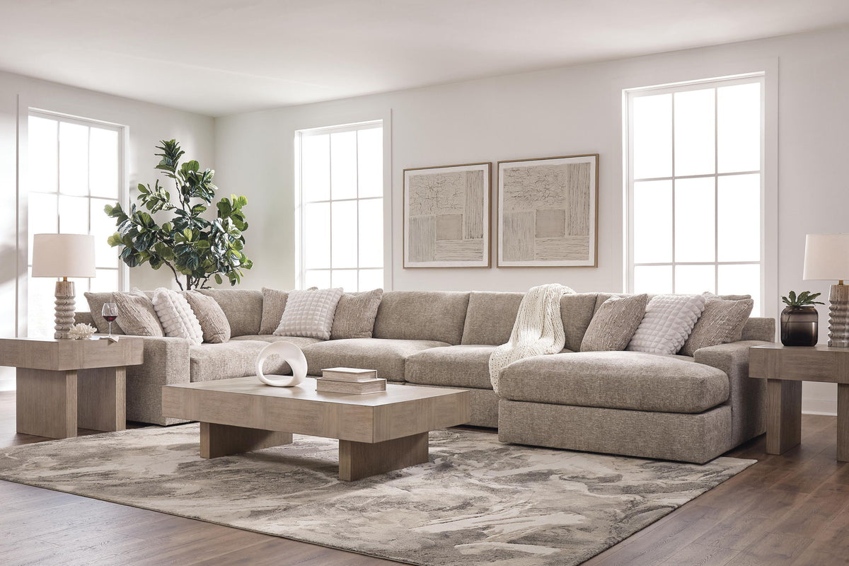 A staged living room featuring a large sectional sofa upholstered in a textured light beige fabric, styled with matching throw pillows, a light wood coffee table, side tables with lamps, a patterned area rug, and a potted plant.