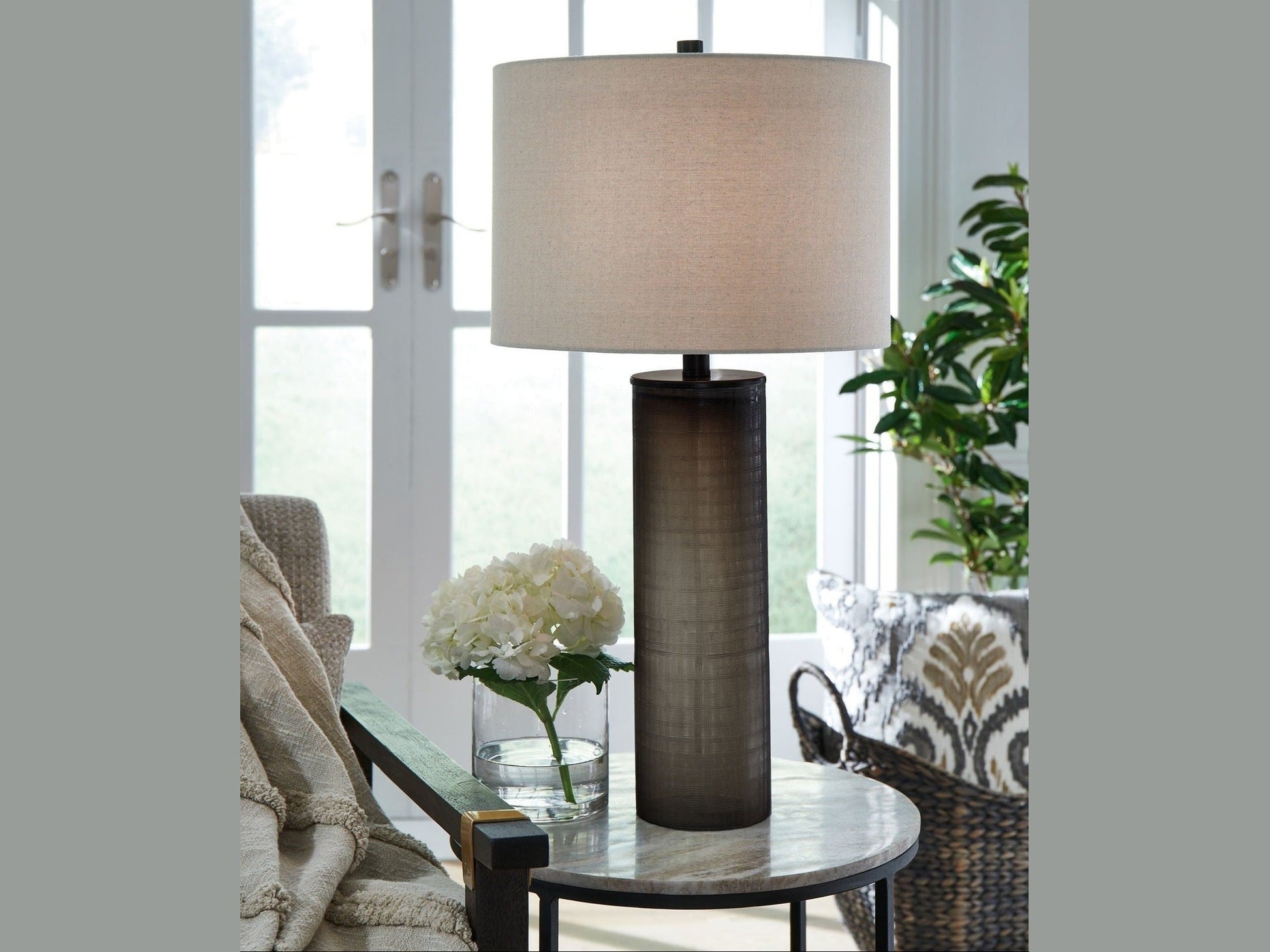 The Dingerly table lamp on a round glass table with flowers and a chair in a bright room.