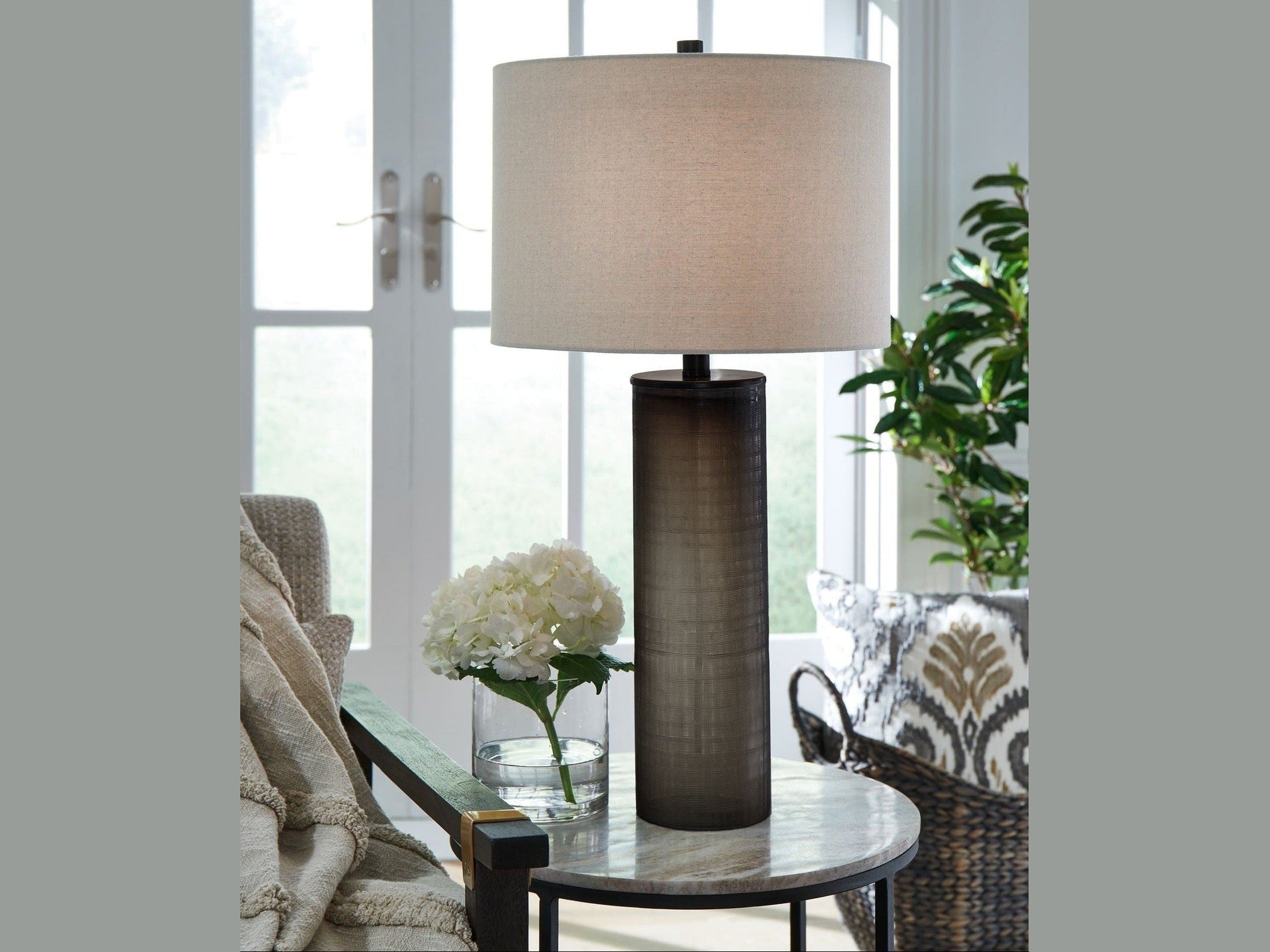 The Dingerly table lamp on a round glass table with flowers and a chair in a bright room.