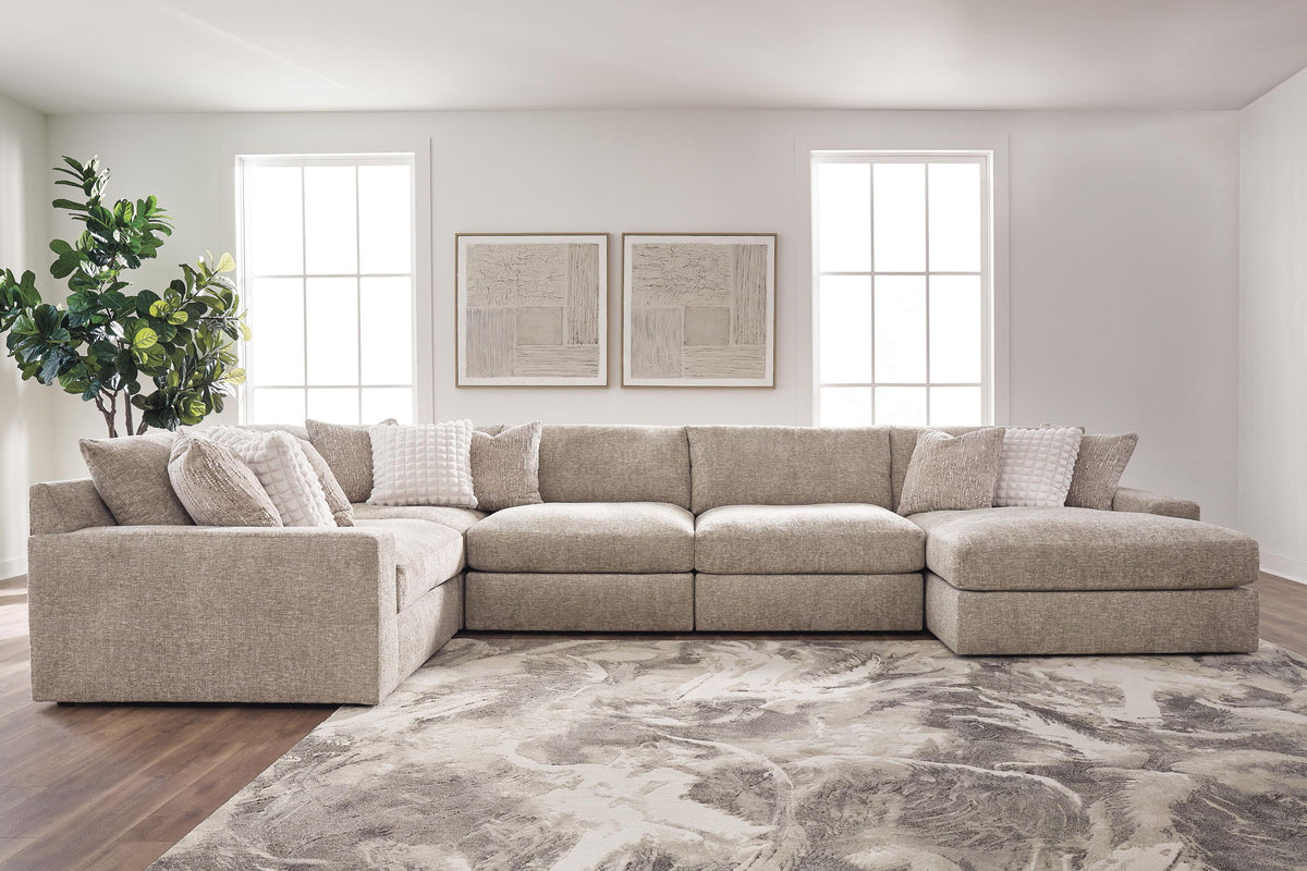 A light beige upholstered sectional sofa with a right-facing chaise lounge, styled in a bright, modern living room with a gray and white patterned rug, dark wood floors, and two large windows.