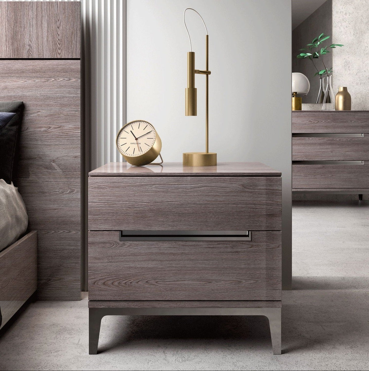 A modern two-drawer nightstand in a gray wood-grain finish with slim metal handles and tapered legs, styled in a bedroom setting with a gold floor lamp and a small round clock on top.