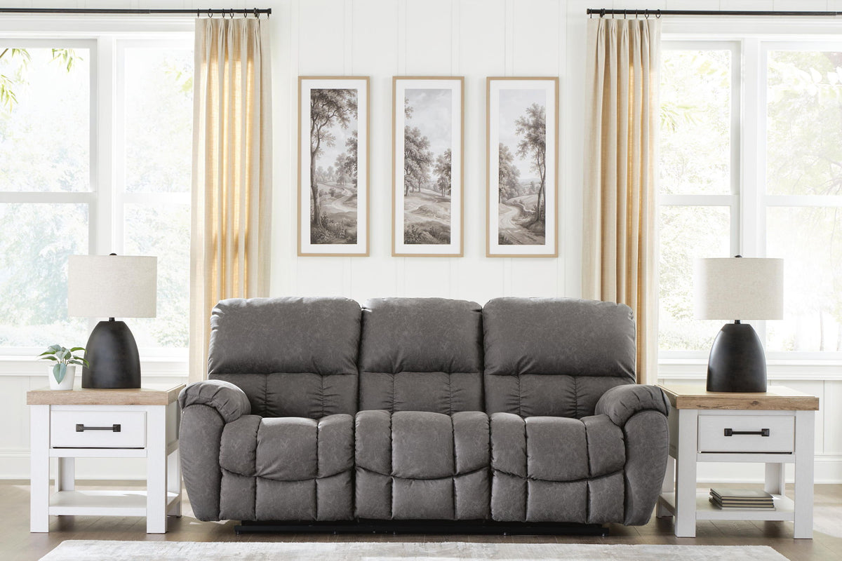 A gray upholstered reclining loveseat centered in a bright, modern living room with white walls, beige curtains, two white side tables with black lamps, and three framed landscape prints above the sofa.