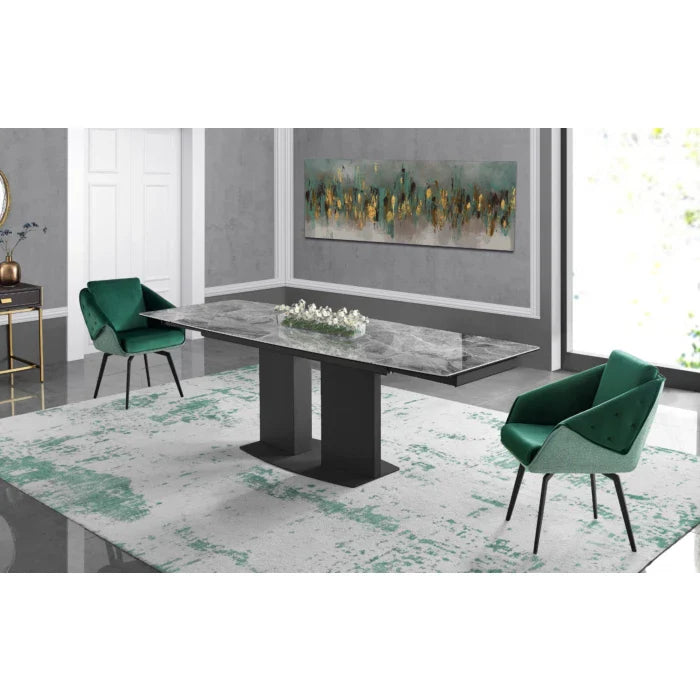 A contemporary grey marble dining table with a black pedestal base sits between two emerald green velvet armchairs on a matching patterned area rug in a modern room.