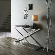 A modern console table with a geometric chrome X-base and black top sits in a minimalist room decorated with a table lamp and wall art.
